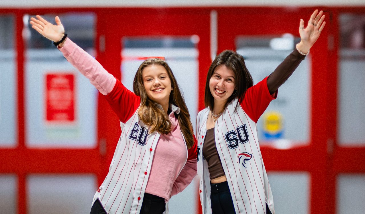 Two young women wearing Brock University Students’ Union shirts pose for a photo each holding an arm up in the air.