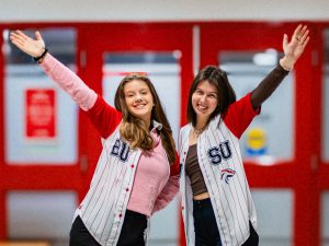 Two young women wearing Brock University Students’ Union shirts pose for a photo each holding an arm up in the air.