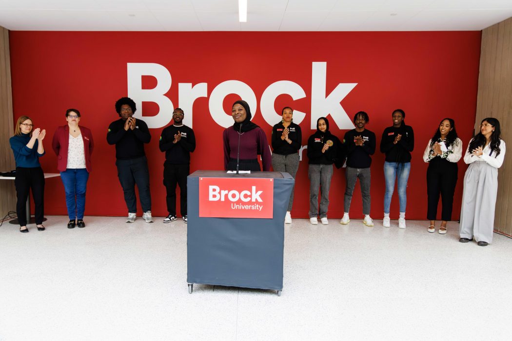 A group of people stand against a red wall with large white letters reading "Brock." A person stands behind a podium in the foreground.
