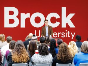 A man speaks to an audience in front of a red wall with the Brock University logo on it.