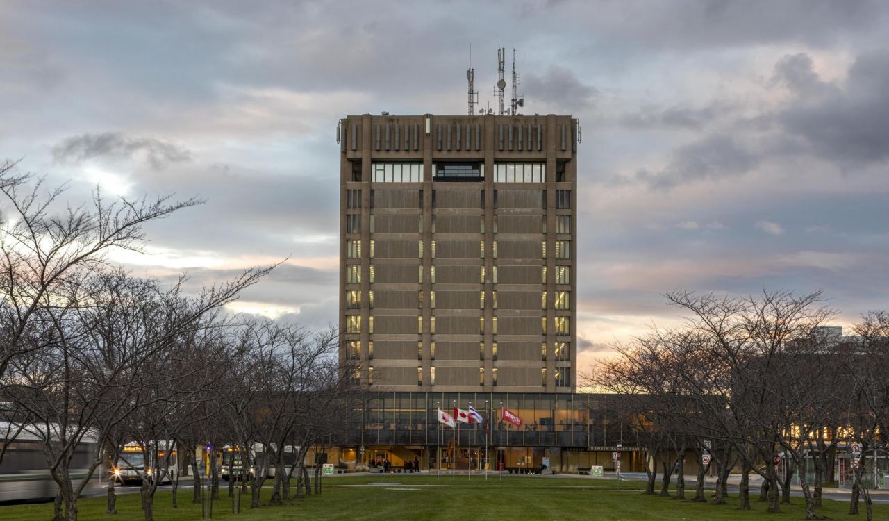 The Brock University Schmon Tower with a sunset in the background and leafless trees in the foreground, three transit buses are parked on the left.