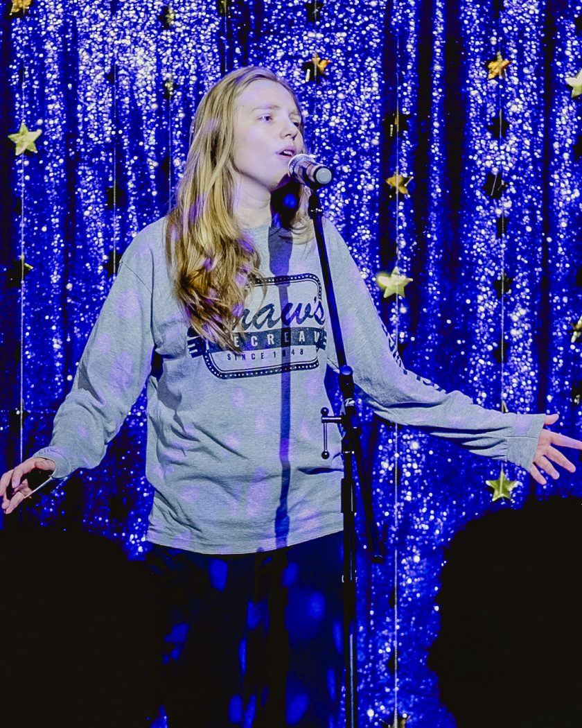 A young woman sings into a microphone on stage in front of a blue backdrop.