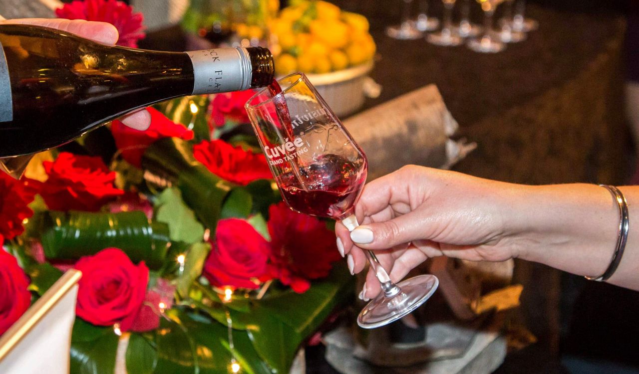 A person pours wine from a bottle into a glass being held by another person. The glass says "Cuvée Grand Tasting."