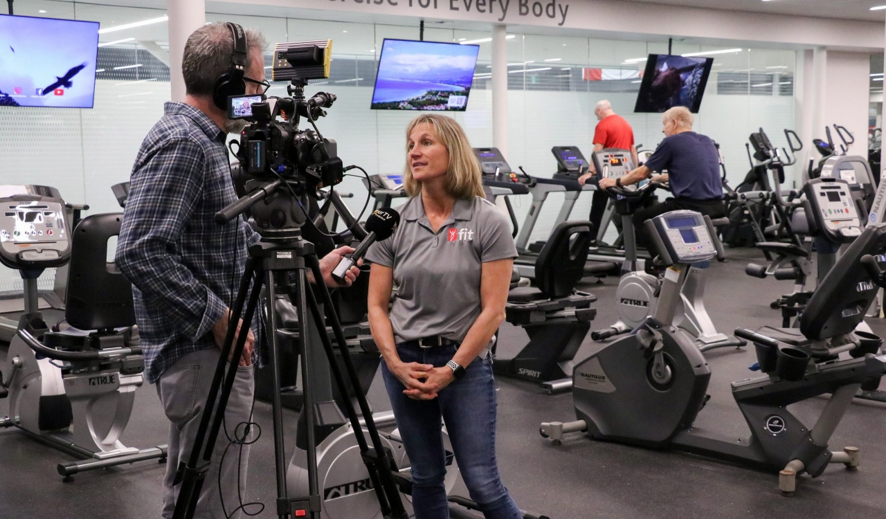 A woman speaks to a camera being held by reporter while standing in front of a gym full of exercise equipment.