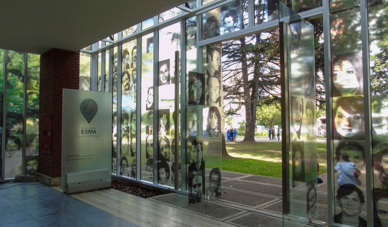 An image from the UNESCO World Heritage Site ESMA Museum and Site of Memory – Former Clandestine Center of Detention, Torture and Extermination shows a glass wall with photos of disappeared people. The lobby door is open and looks out onto a green field with people walking outside on a sunny day.