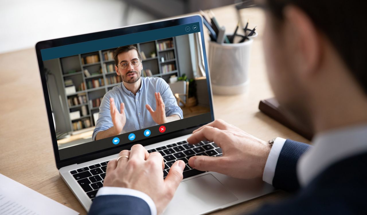 A businessman, seen from behind, talks with another man on a video call using a laptop.