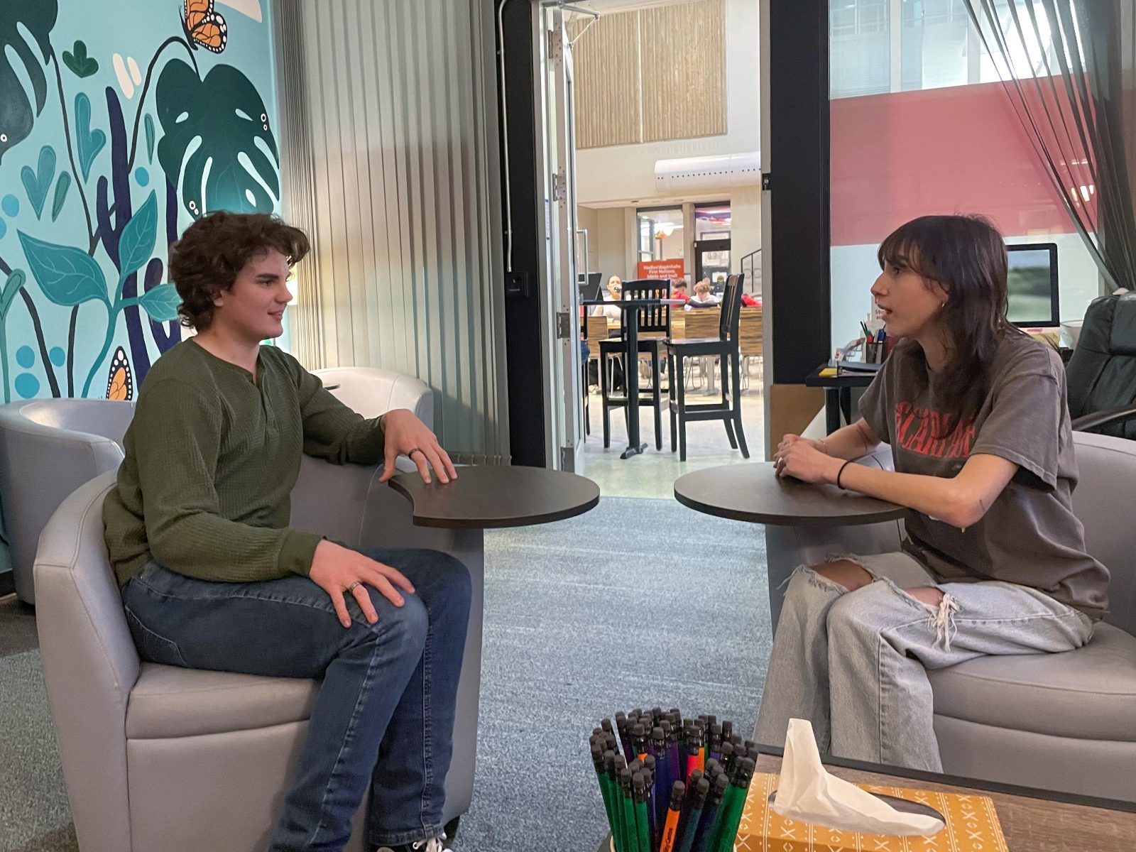 Two university students sit in armchairs having a conversation. A busy cafeteria can be seen through the open door behind them.