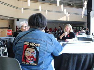 A woman wears a jacket with ‘no more stolen sisters’ messaging on it while speaking with two other women at the table.