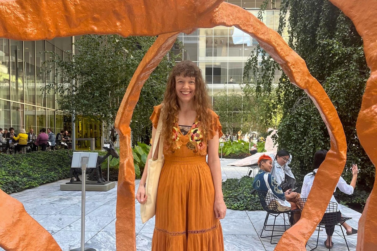 Jackie Armstrong stands in New York City in a courtyard beneath a sculpture. She is smiling warmly wearing a bright orange dress.