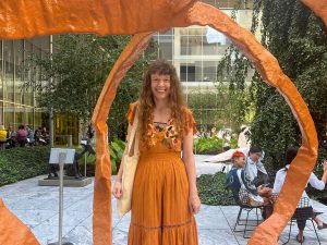 Jackie Armstrong stands in New York City in a courtyard beneath a sculpture. She is smiling warmly wearing a bright orange dress.