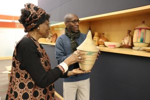A woman in a library hands a man a woven jar from Rwanda. Behind them is a shelf holding similar objects.