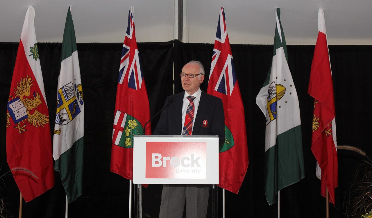 A man stands at a podium with a series of flags in the background.