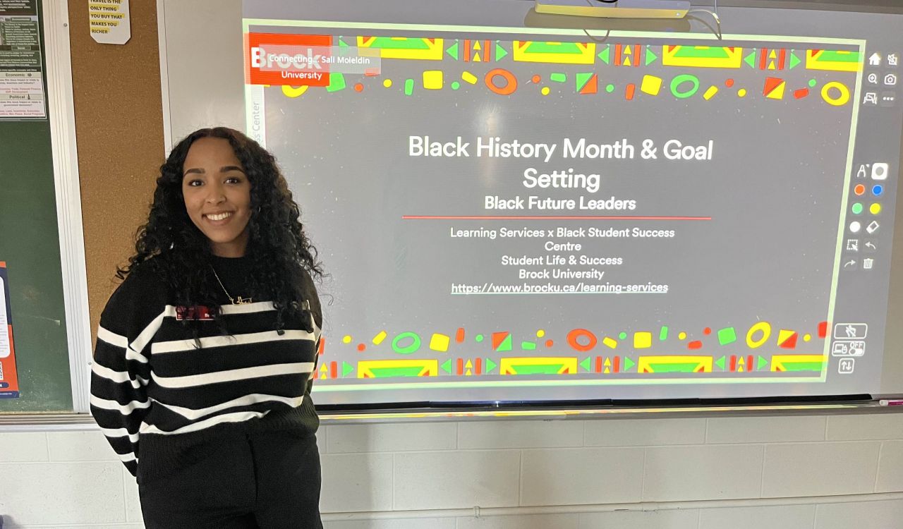 A woman stands ready to give a presentation on Black History Month at the front of a high school classroom.