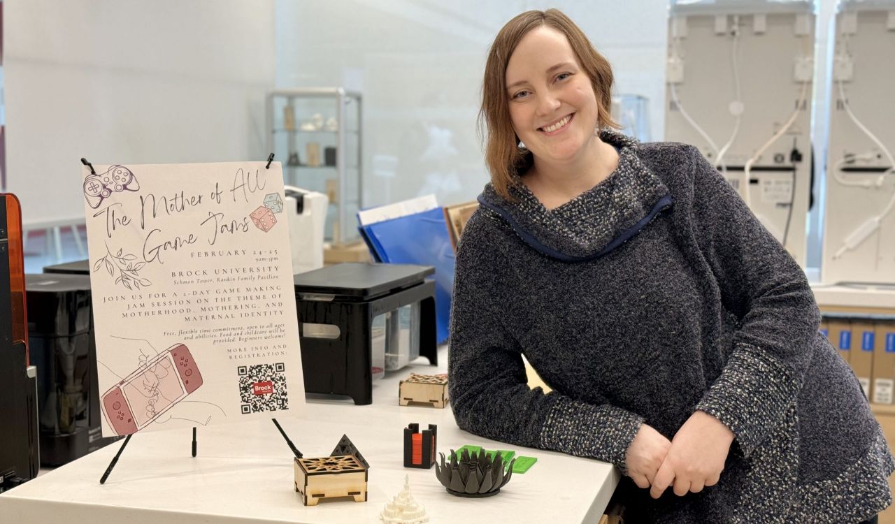 Brock Assistant Professor Sarah Stang stands in the Brock Library Makerspace with 3D printing technology in the background. She leans against a table showing an event poster and small 3D printed items that can be used for board games like small circles and a flower, and box.