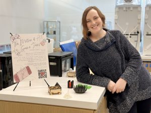 Brock Assistant Professor Sarah Stang stands in the Brock Library Makerspace with 3D printing technology in the background. She leans against a table showing an event poster and small 3D printed items that can be used for board games like small circles and a flower, and box.
