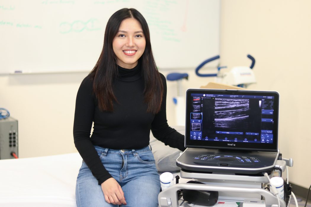 Shamae Quinquito in an Applied Health Sciences lab at Brock University. She is standing/sitting on a bed next to an ultrasound machine.