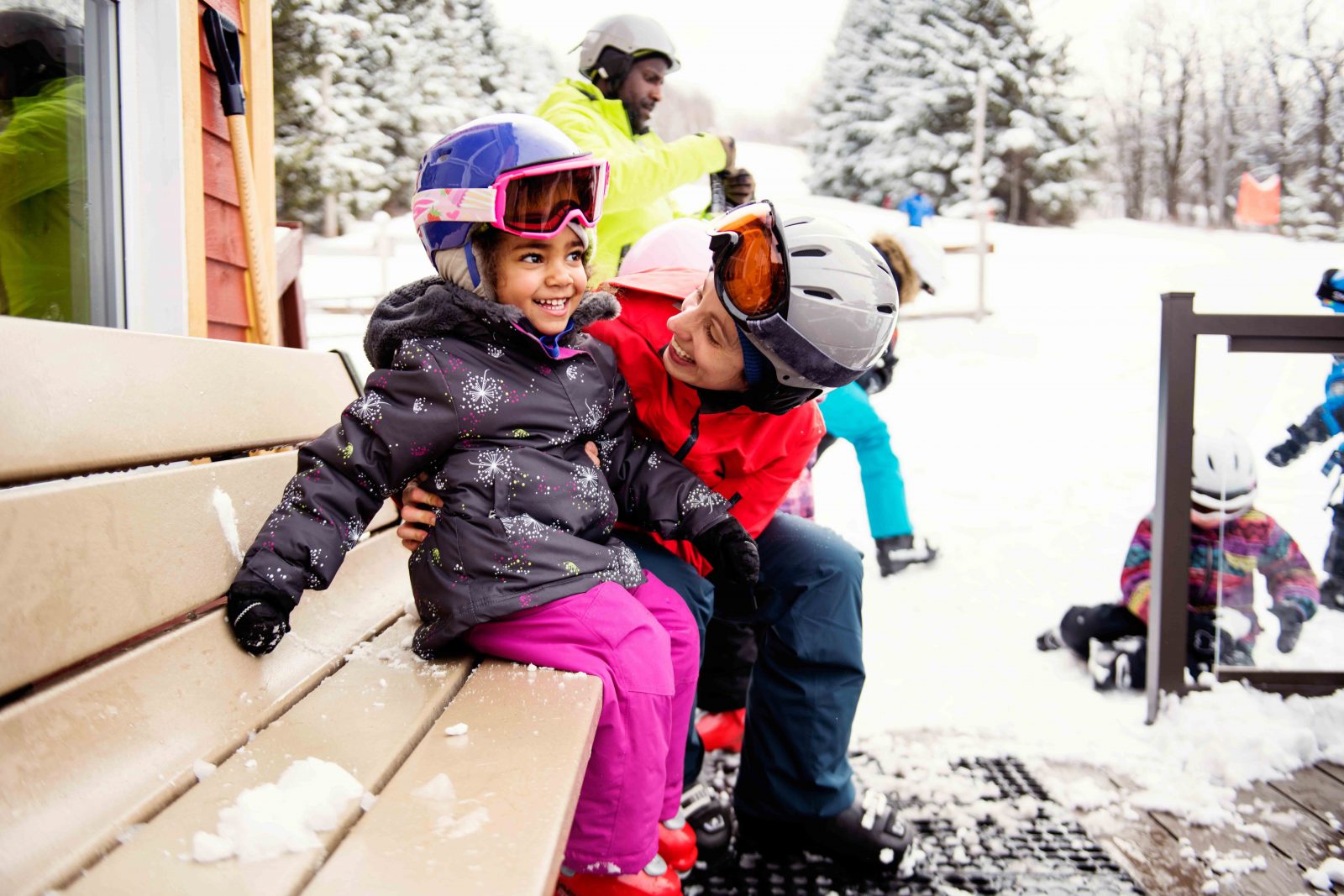 A mother and child sit on a bench preparing to go skiing.