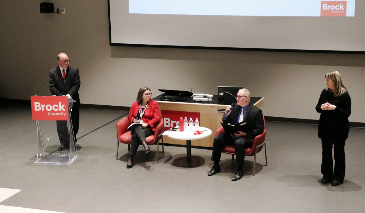 Four people engage in discussion at the front of an auditorium, two seated and one at a podium.