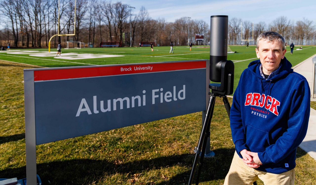 Thad Harroun stands next to a sign for Alumni Field. Behind him are several athletes playing soccer on an outdoor turf field. He is wearing a Brock University sweater, and next to him is a large telescope.