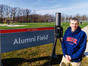Thad Harroun stands next to a sign for Alumni Field. Behind him are several athletes playing soccer on an outdoor turf field. He is wearing a Brock University sweater, and next to him is a large telescope.