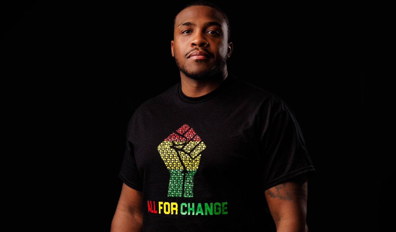 A man stands in the dark wearing an inclusive T-shirt in honour of Black History Month and African Heritage Month.