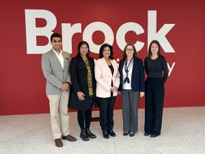 Five people pose side-by-side in front of a red wall with the Brock University logo on it.
