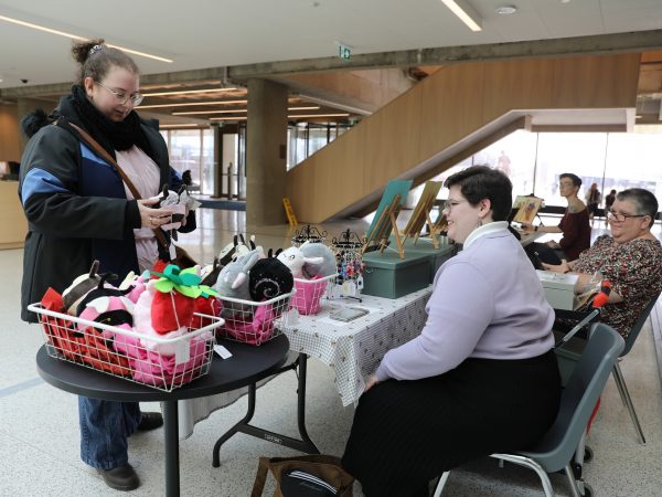 A woman holding a plush toy while standing beside a table of crafts with two women seated on the other side.