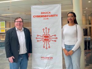 A man and a woman pose next to a Brock Cybersecurity Club banner.
