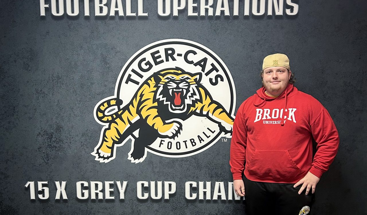 Alex Penz, wearing a red Brock University hoodie sweater, stands in front of a wall sign with the Hamilton Tiger-Cats logo that reads “Football Operations. 15-time Grey Cup Champions.”