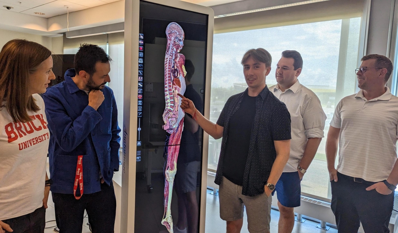 A man points at a picture of a human body on a large virtual screen while surrounded by his colleagues.