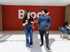 Two students walk in a sunny atrium in front of a red Brock wall.