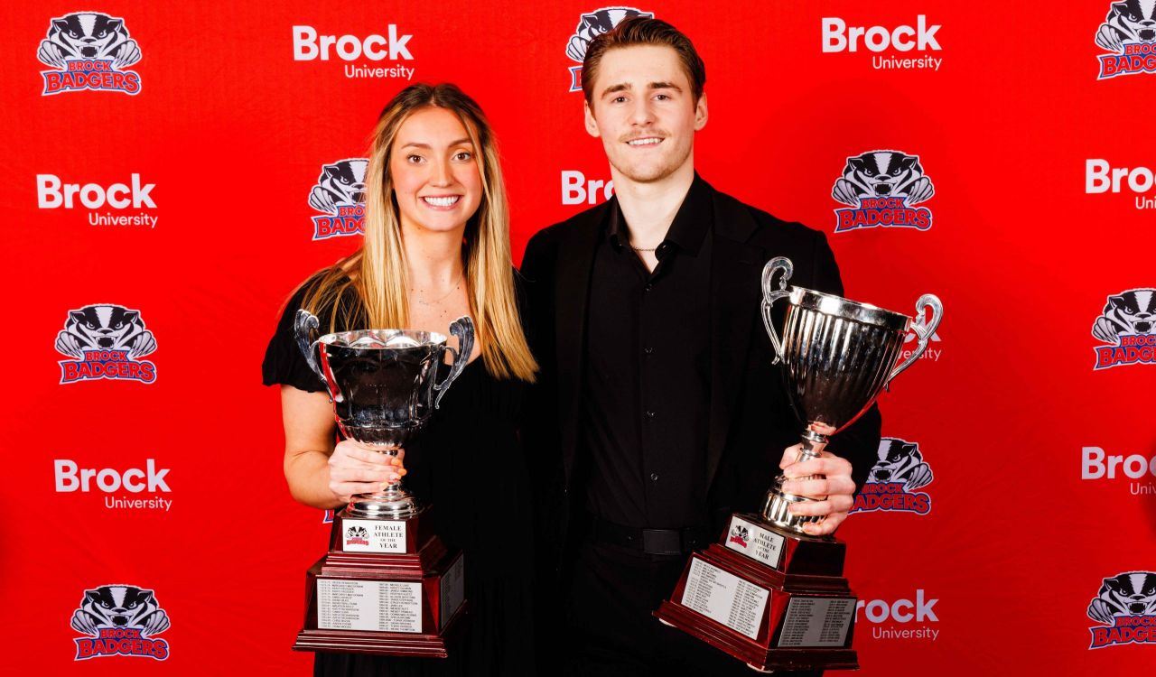 A woman and a man stand side by side holding trophies in front of a Brock University-branded backdrop.