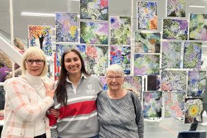 Three women stand next to each other in front of a colourful art display on a clear wall. The two women on the end are seniors. The woman in the middle is in her 20s.