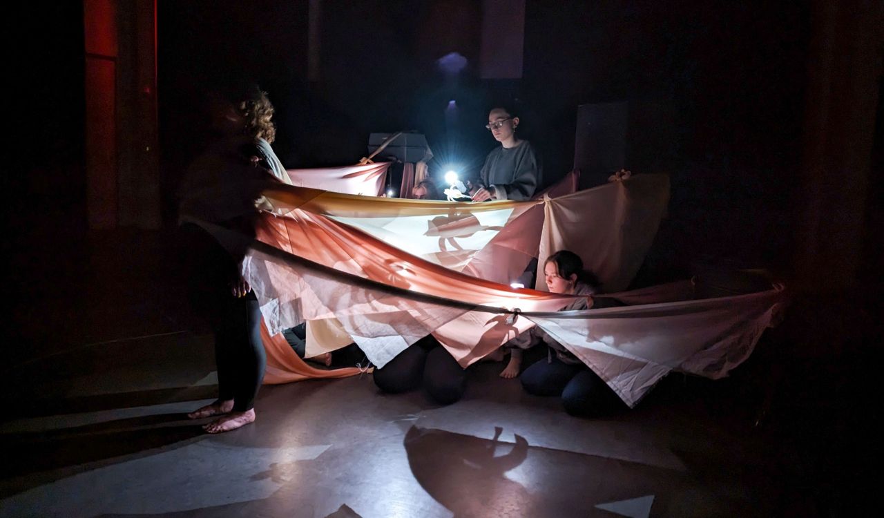 A dark image with shadows show Dramatic Arts students rehearsing on a stage with fabric and shadow puppets. Lighting effects are used to create bright pockets of colour behind the material held up by students cloaked in darkness while shadows in geometric shapes are cast on the stage floor.