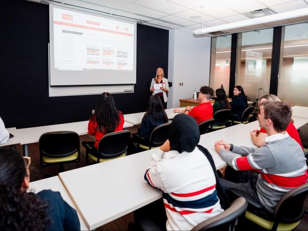 A group of students in a university classroom, seen from behind, look at an instructor in front of a presentation screen.