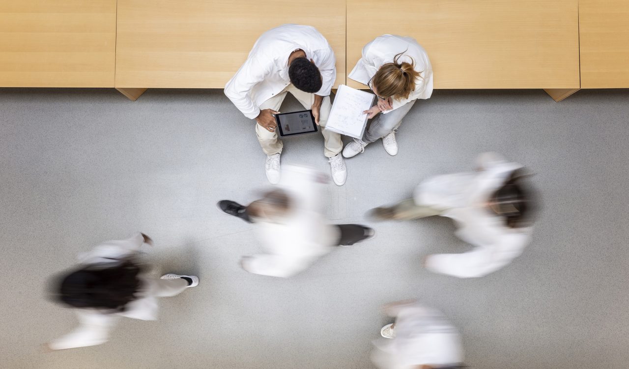 Two people in white lab coats sit on a bench looking at a tablet and clipboard. Blurry, out-of-focus people in lab coats walk past them.