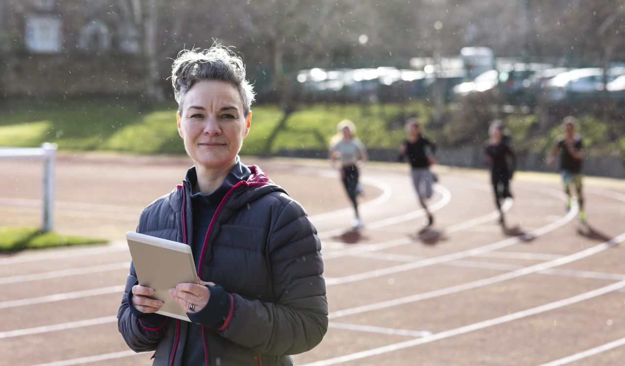 A shot of a female sports coach standing and looking into the camera. There are female athletes running past in the background. It is a bright day with slight rain.