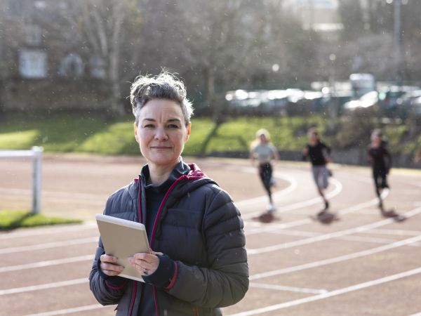 A shot of a female sports coach standing and looking into the camera. There are female athletes running past in the background. It is a bright day with slight rain.