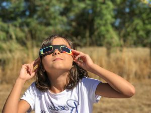 A young girl looking at the sun through eclipse glasses during a solar eclipse at an outdoor event.