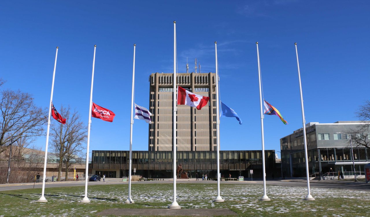 A row of six flags sit at half mast in front of Brock University.
