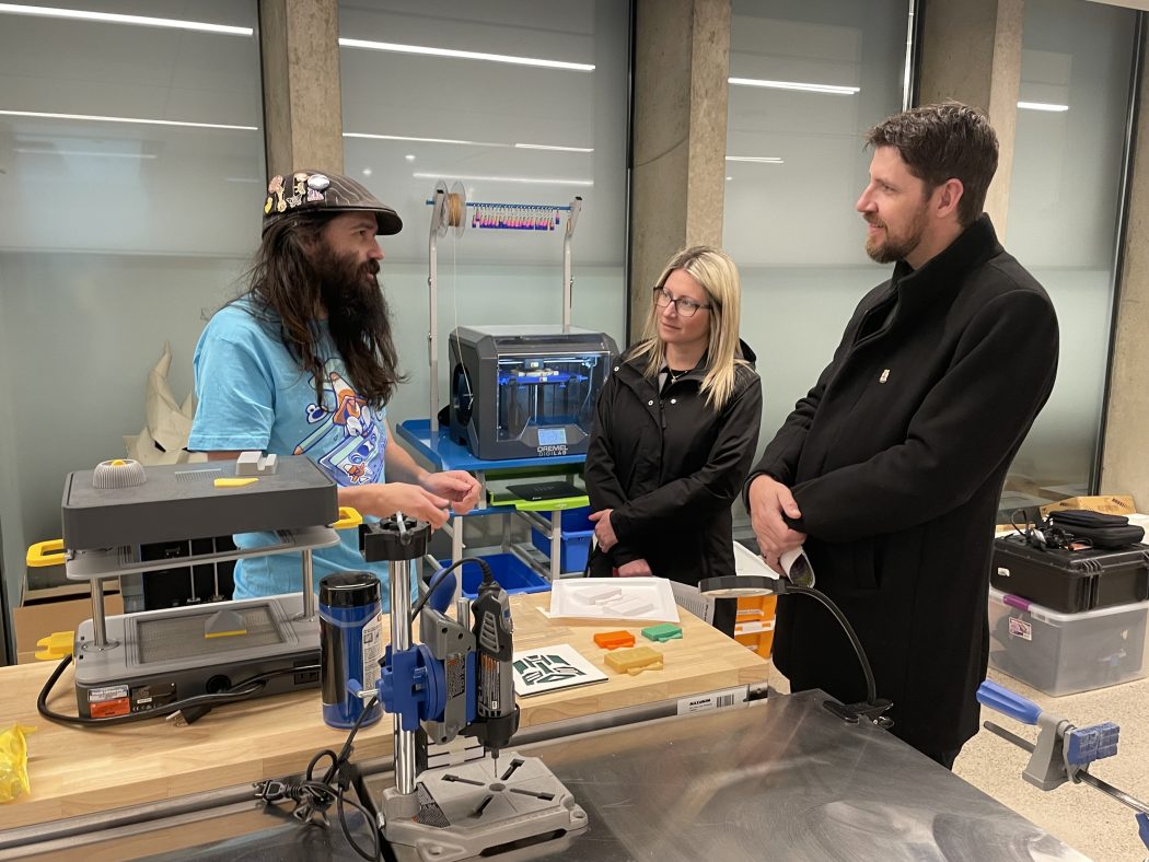 Three people discuss technology beside equipment in a library makerspace.