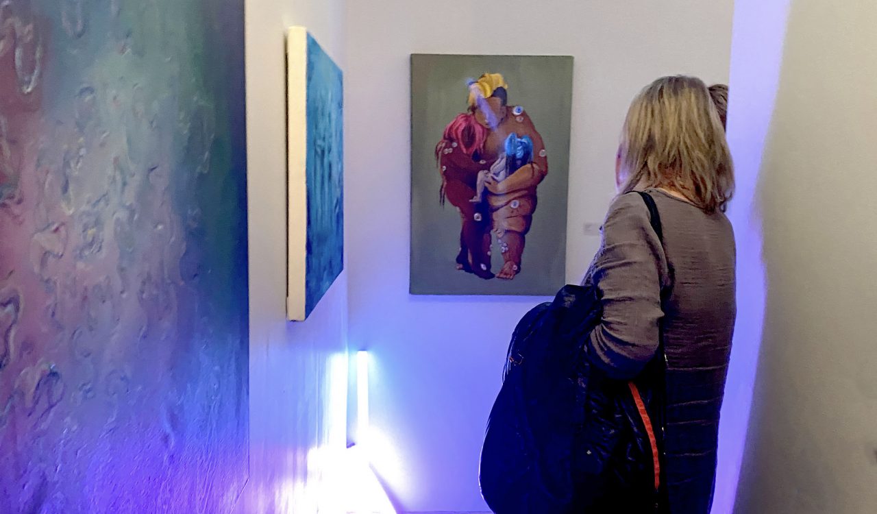 A woman with her back to the viewer looks at a painting hanging on a white wall.
