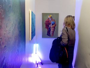 A woman with her back to the viewer looks at a painting hanging on a white wall.