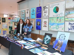 Sarah McGean and Marie Bowering at a booth featuring colourful artwork from the students of Soaring Eagles Indigenous Elementary School the first annual World Water Day Celebration.