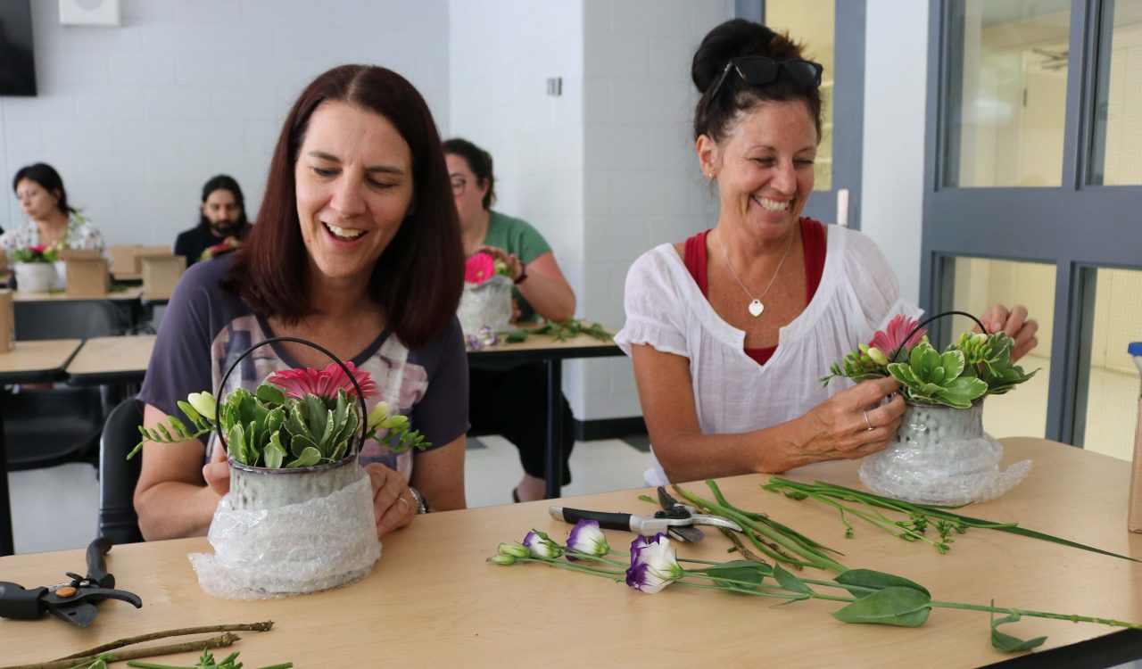 Two women sit at a table assembling floral arrangements in a classroom.