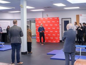 Peter Tiidus stands in front of red Brock University backdrop and speaks to a crowd of people in a large physical activity room.