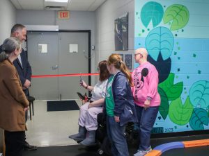 Five people gather near a red ribbon placed horizontally over the opening of a hallway. One of the people cuts it with scissors in celebration of the CAPA facility opening.