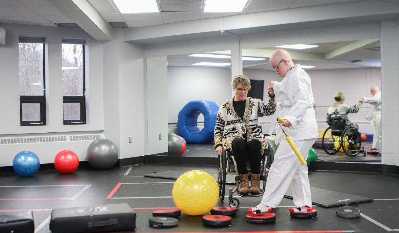 A woman using a wheelchair holds the hand of a woman dressed in a white martial arts robe as she walks across balance boards in a large fitness studio.