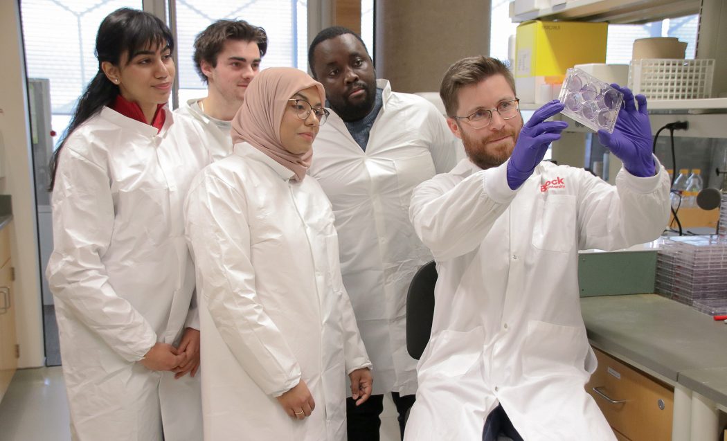 Assistant Professor of Biological Sciences Ian Patterson (far right) sits on a lab chair holding up, and looking at, a clear box with light purple circles in the middle, while four students all in white lab coats standing behind him look at the box. 