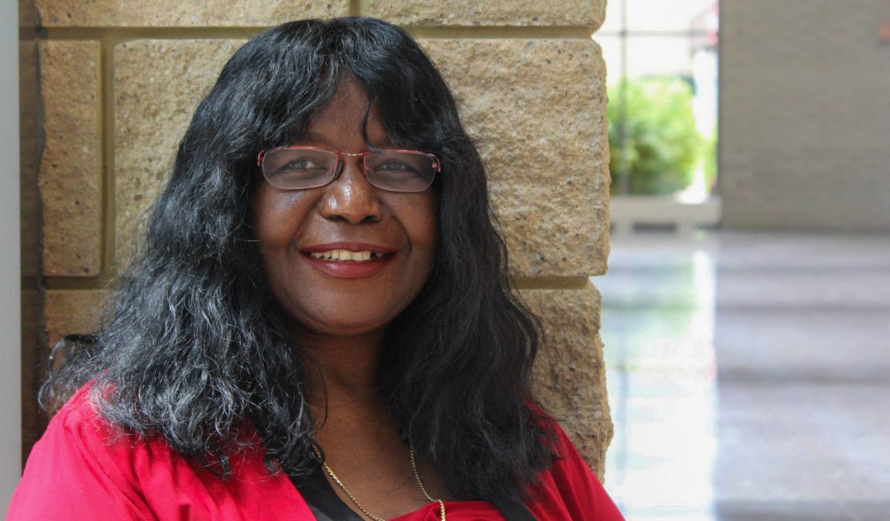 A portrait photo shows Brock Professor Joyce Mgombelo standing in a bright hallway, leaning against a beige wall. Shown from the shoulders up, she wears a red shirt and red glasses, smiling warmly at the camera.